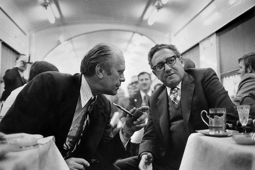 A black and white photo of two men on an aeroplane, leaning across the aisle to talk. 