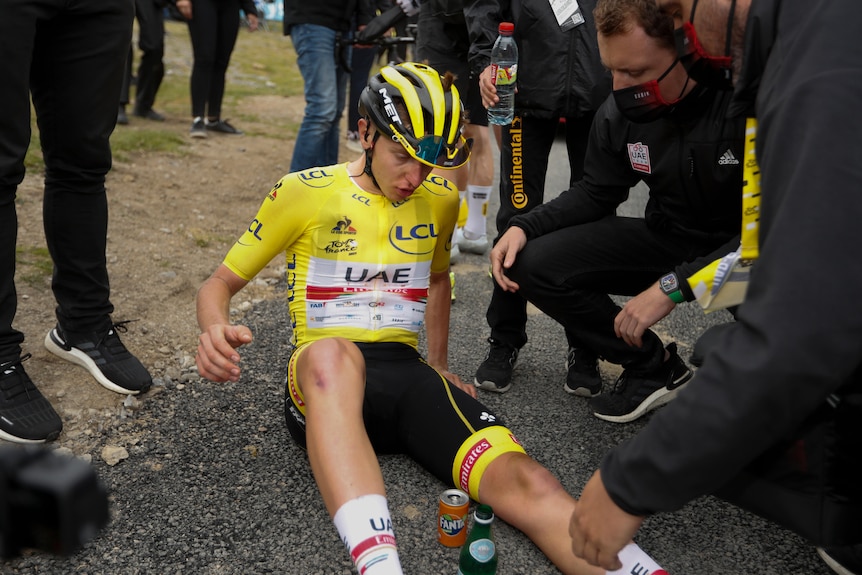 An exhausted cyclist wearing a yellow jersey sits on the ground looking at his leg while team officials check on him.