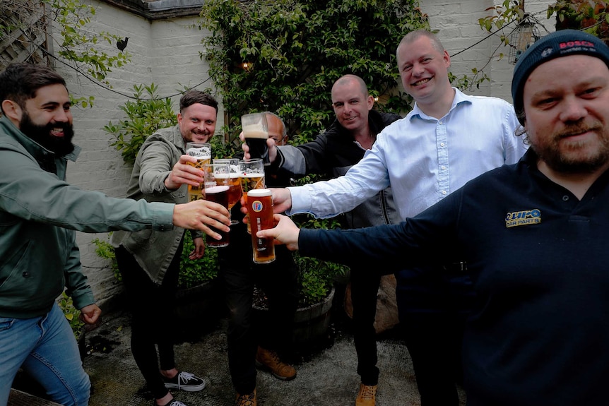 A group of men clink beer glasses and smile as they stand in an outdoors area.