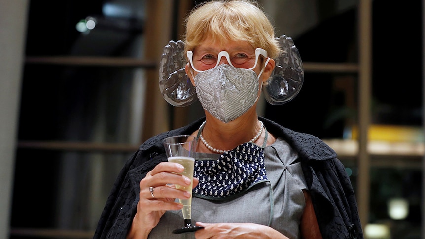 A woman wears a face mask with transparent plastic hands cupped around the ears. She holds a glass of champagne.