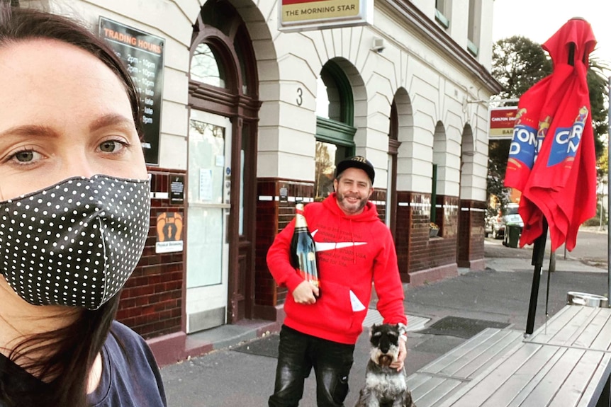 A woman wearing a mask and man in a red hoodie holding a large bottle of champagne outside a pub on a grey day.