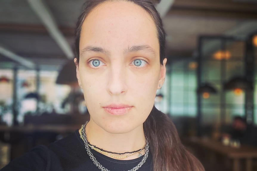 A young woman with pale blue eyes stares just off camera, wearing black Tshirt and two necklaces