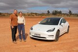 A man and a woman stand next to a white car with red desert dirt stretching behind them