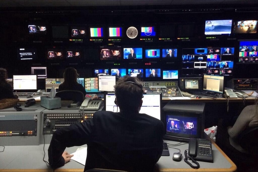 Back of man sitting in chair watching a bank of TV monitors in a studio control room.