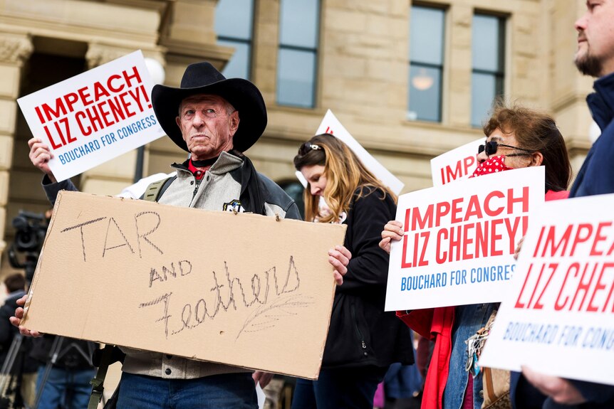 A man wearing a Stetson carries signs saying IMPEACH LIZ CHENEY and TAR AND FEATHERS in a protest crowd