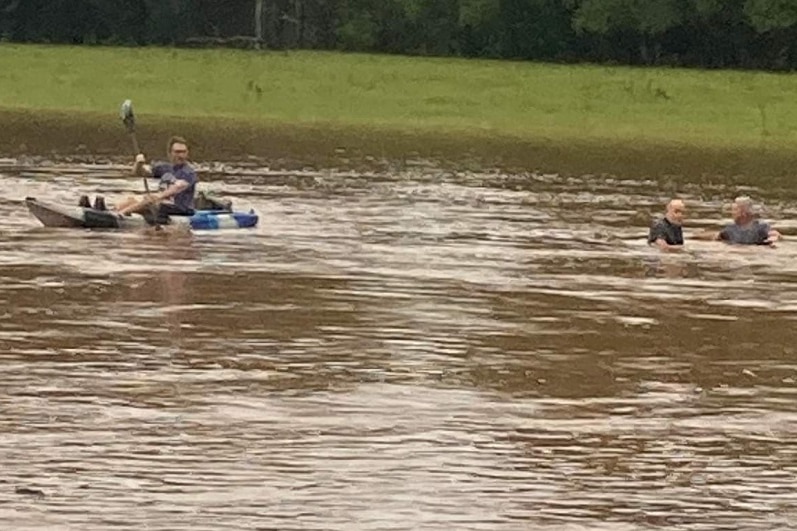 Two men standing on the back of a ute while another paddles to them on a kayak.