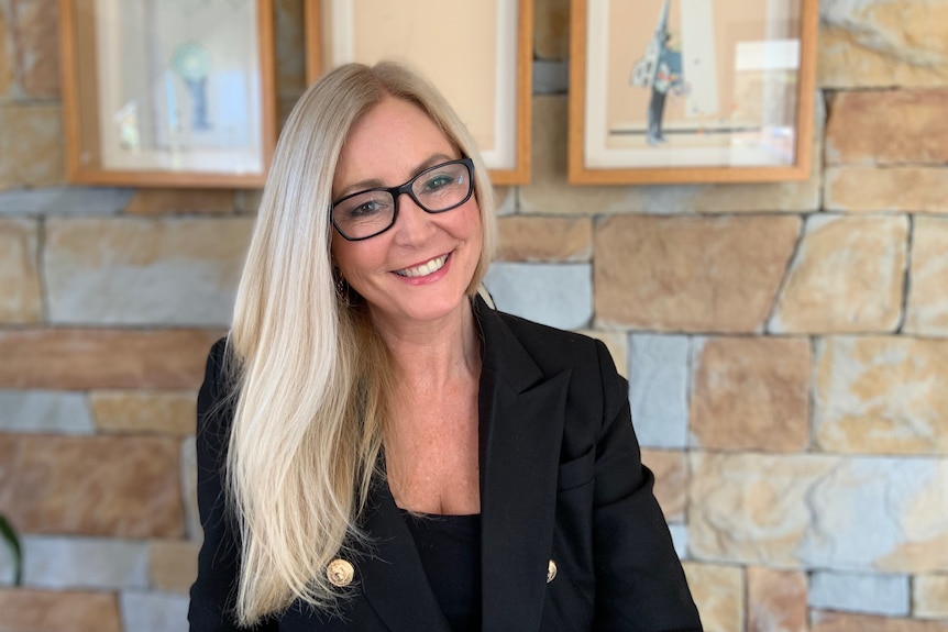 A woman wearing a black blazer and glasses sitting in front of a stone wall smiles for a photo.