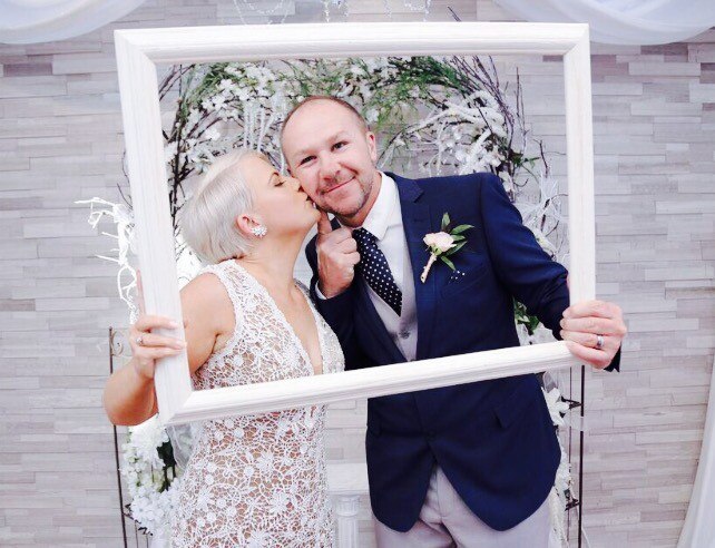 Photo of a woman and man holding up a photo frame and kissing each other on the cheek in a chapel