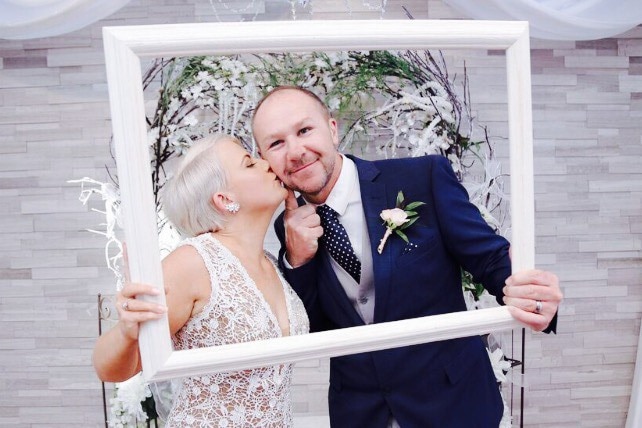 Photo of a woman and man holding up a photo frame and kissing each other on the cheek in a chapel