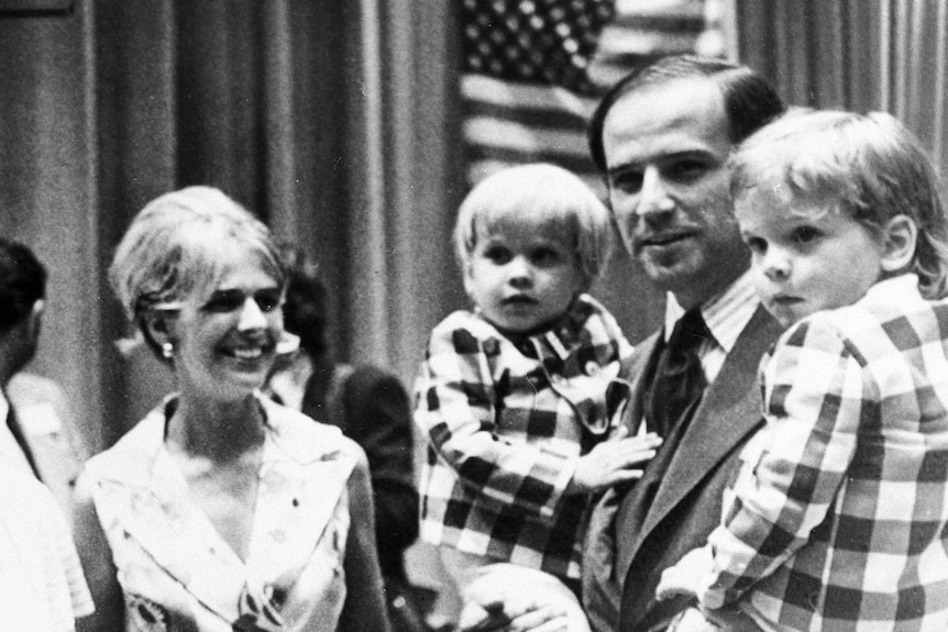 Black and white photo of joe biden holding his two young sons on his hip and standing next to his late wife