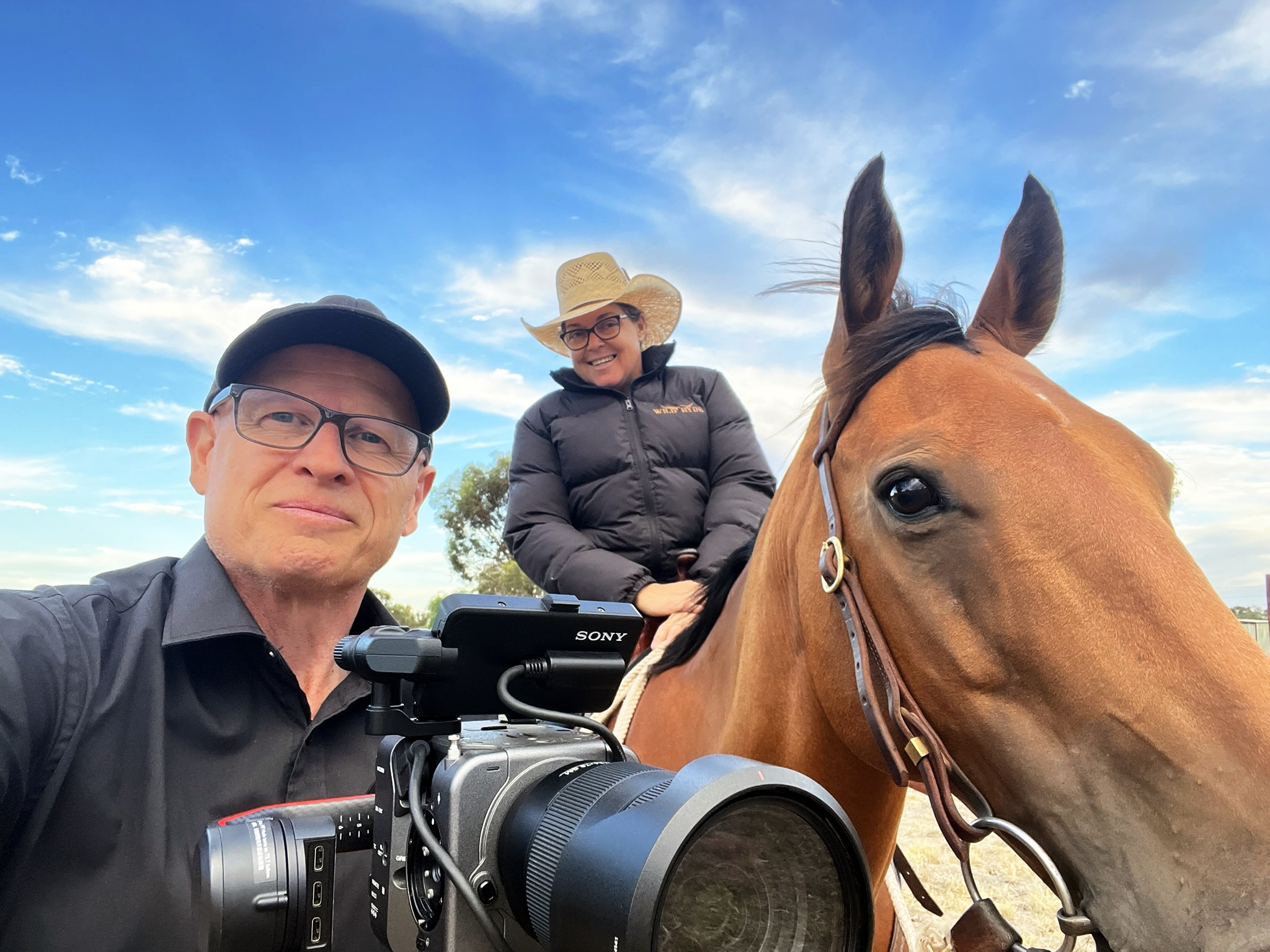 A close-up selfie of a woman on a horse in a cowboy hat and a man holding a large digital camera. 