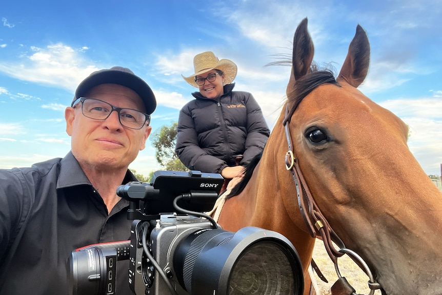 A close-up selfie of a woman on a horse in a cowboy hat and a man holding a large digital camera. 