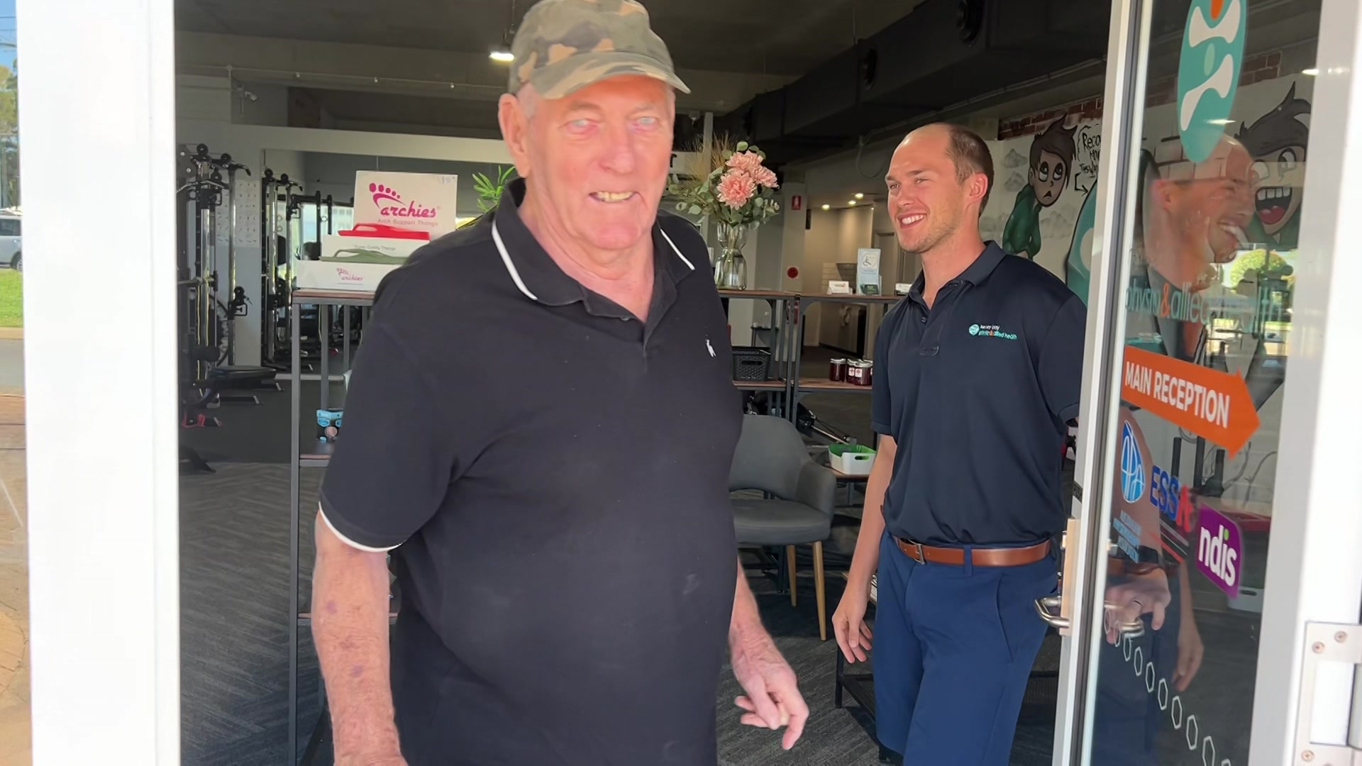 Um homem idoso saindo de uma academia com um homem mais jovem segurando a porta aberta.