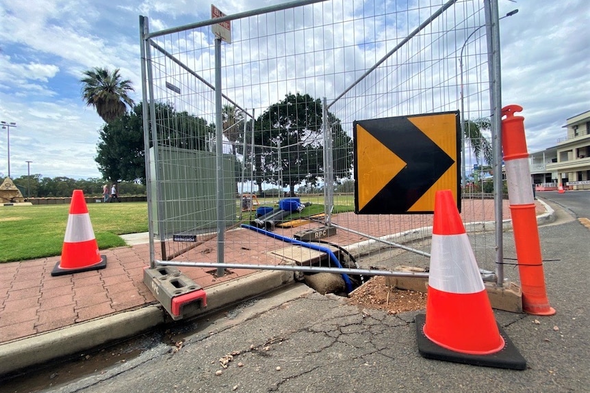 A bituminous road, with bright orange cones, and a metal fence, with a hose leading into the river.