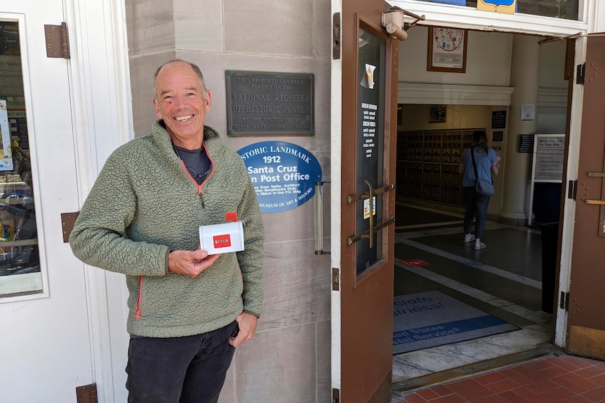 A smiling man holds a Netflix branded letterbox outside the entrance to a post office.