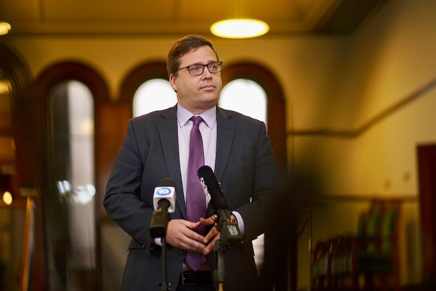 A man in a suit, purple shirt and purple tie and black glasses stands in front of media microphones