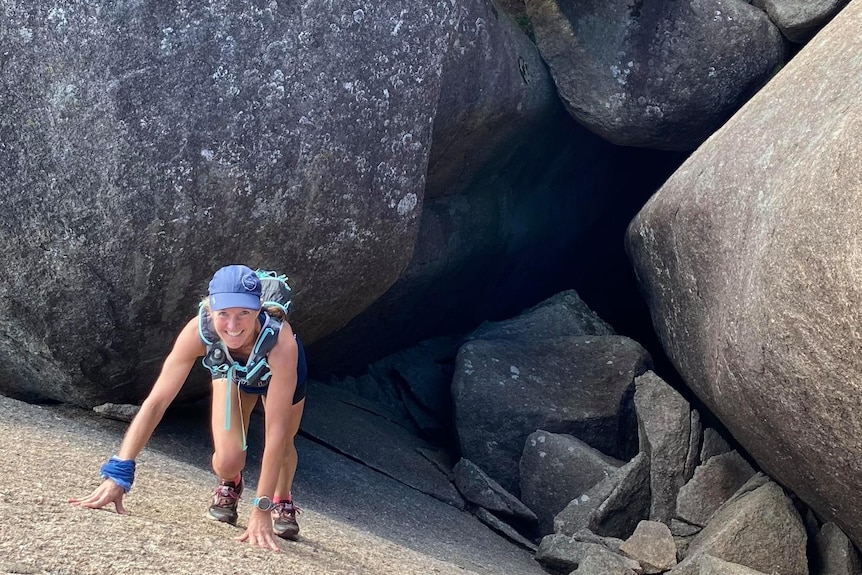 Shiree smiles while looking up, climbing a giant rock, wearing a hat.