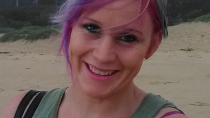 A smiling woman in her 30s, standing on a beach in front of a Norfolk pine. She has purple hair.