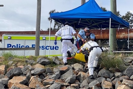 Two men in white  jumpsuits carry something up the riverbank of stones into a small blue marquee as police help.