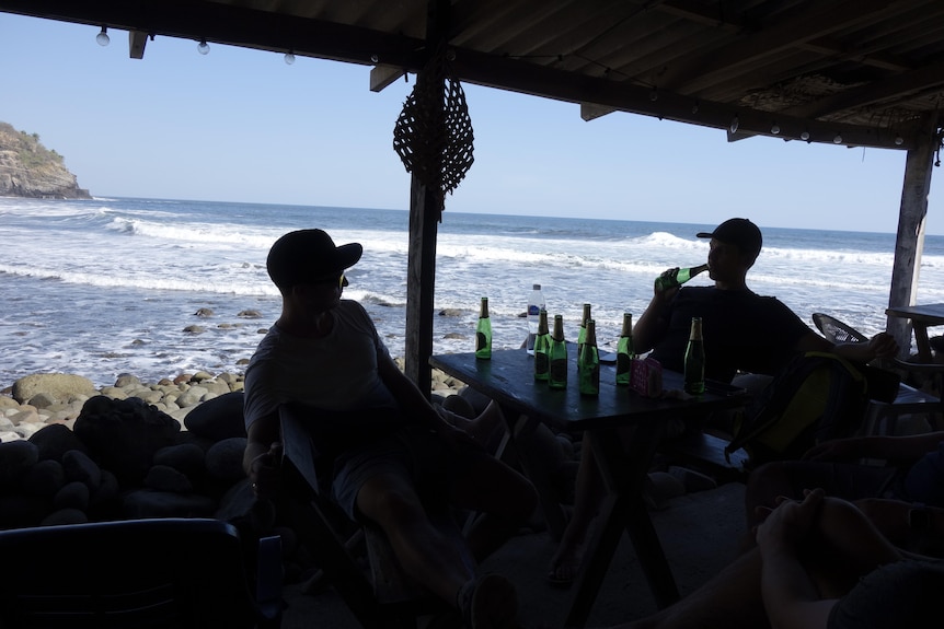 Two men sit at a table drinking beer with the ocean behind them.