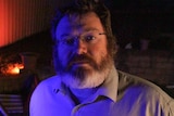A middle-aged man with a beard and spectacles in a dark room.