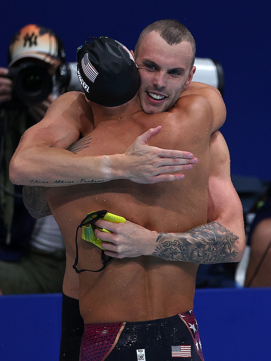 A man hugs another man wearing a black swimming cap