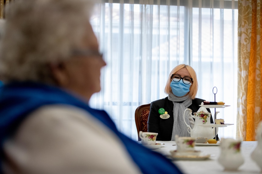 A woman in a mask sits a table with elderly woman drinking tea.