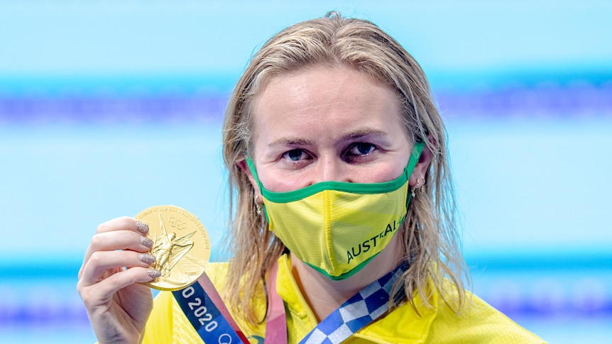 A blonde woman wearing a yellow face mask holds a gold medal