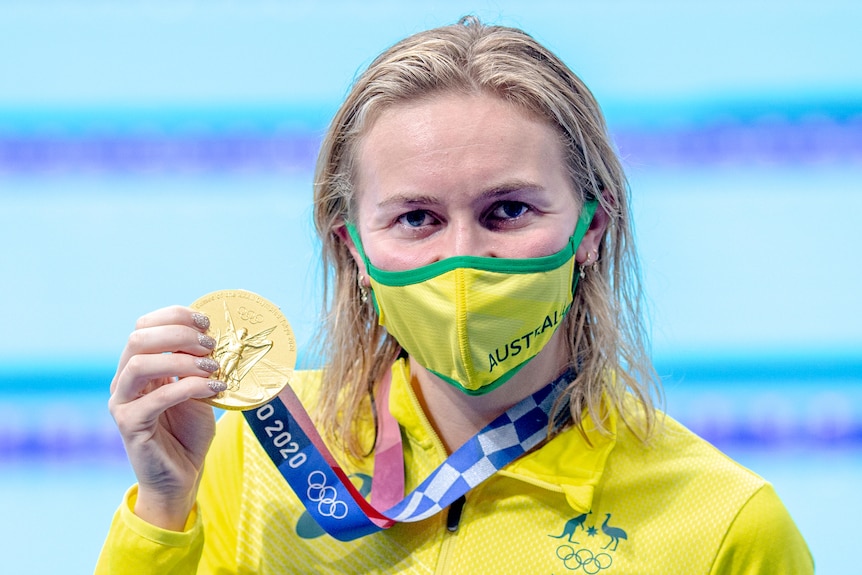 A blonde woman wearing a yellow face mask holds a gold medal