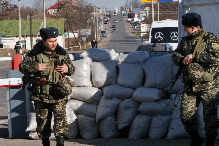 Two men in green camouflage uniforms and dark winter hats stand behind a sandbag pile on a paved road.