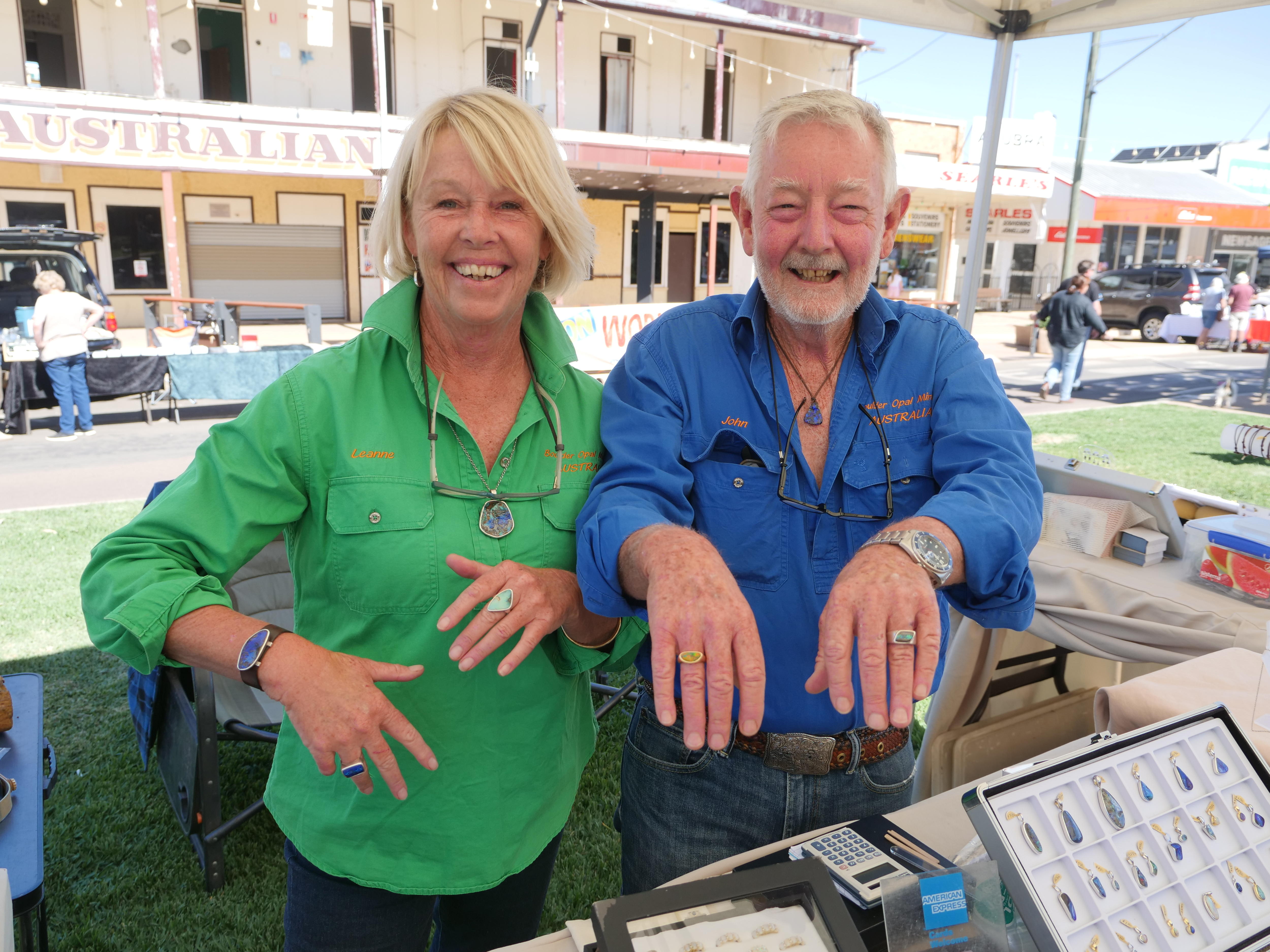 A smiling blonde woman and an older, bearded man smile while showing off their rings at an open-air stall.