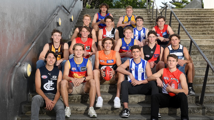 A group of young Australian Rules footballers sit on the steps outside a building wearing the guernseys of their new AFL clubs.