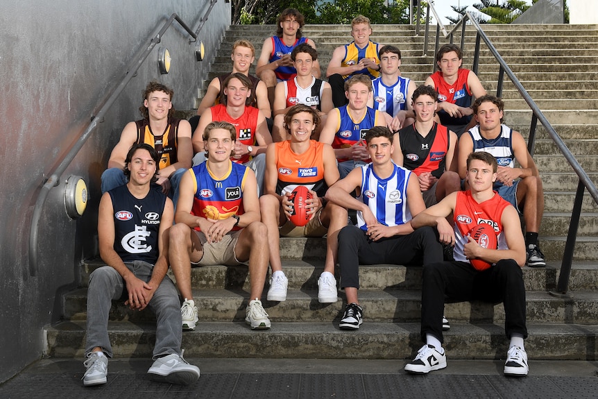 A group of young Australian Rules footballers sit on the steps outside a building wearing the guernseys of their new AFL clubs.