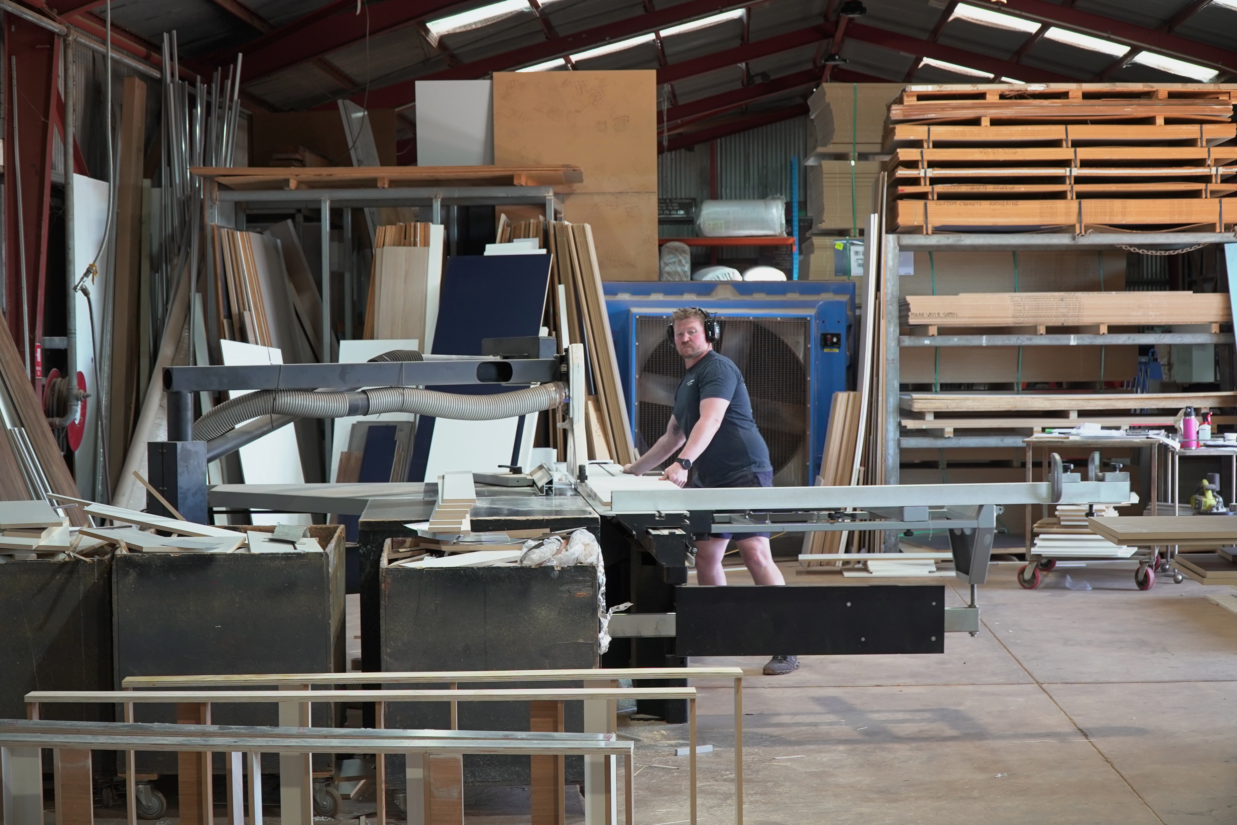 A man standing in a workshop with wood samples around him, slicing wood on a saw.