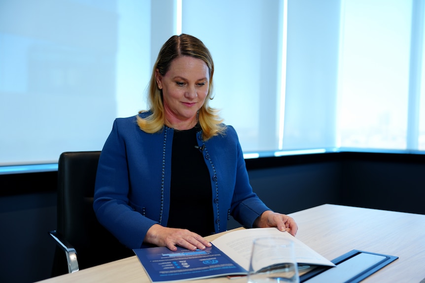 A woman sits at a table looking at a booklet. She wears a blue jacket of a black top and has fair hair.