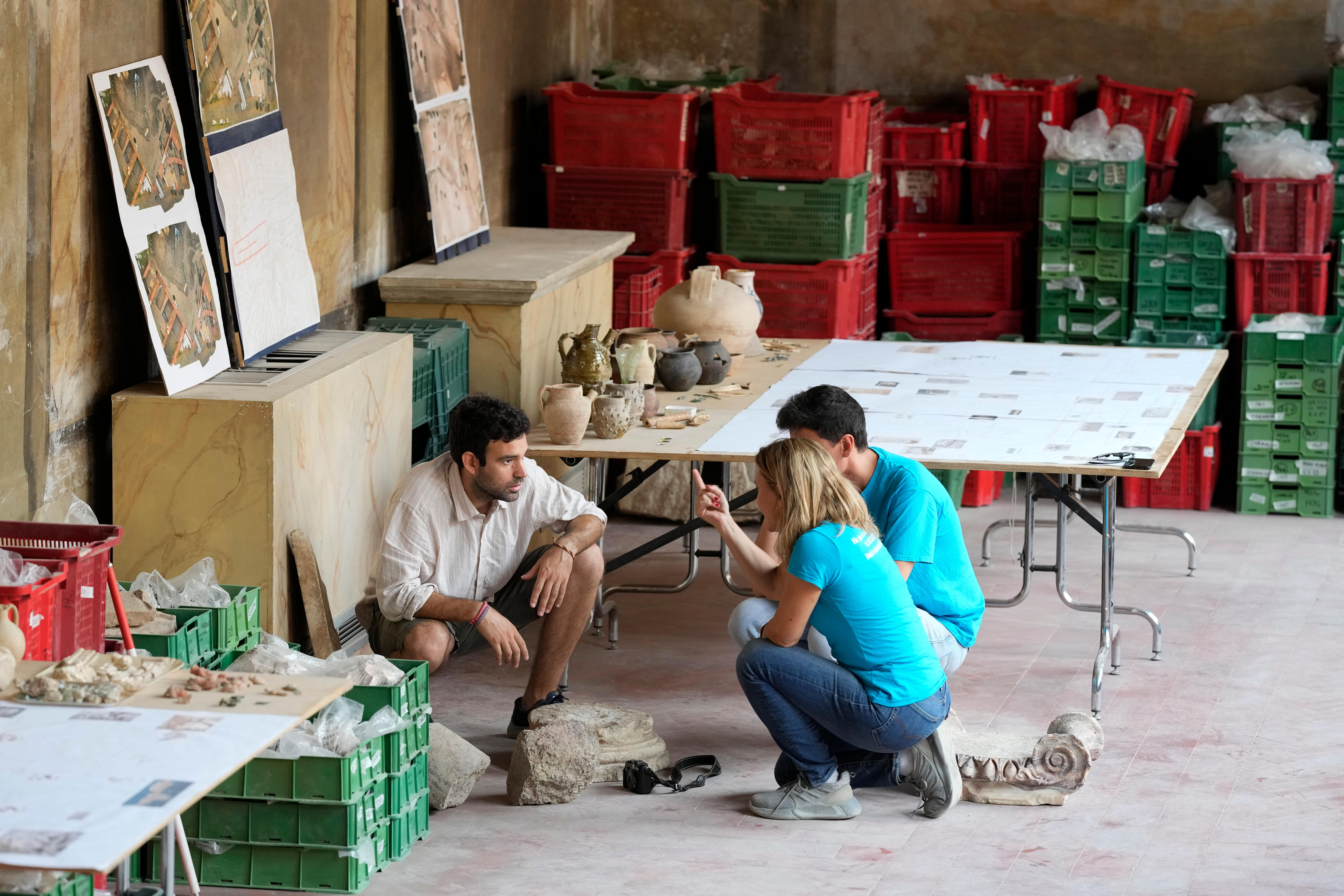 Three people kneeling over some roman era ruins surrounded by a room full of artifacts