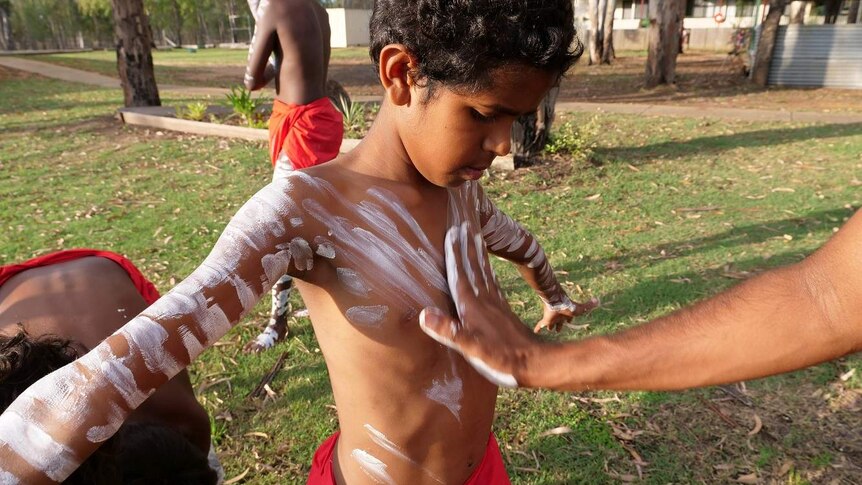 An boy in a traditional indigenous outfit in gets white paint painted on his chest