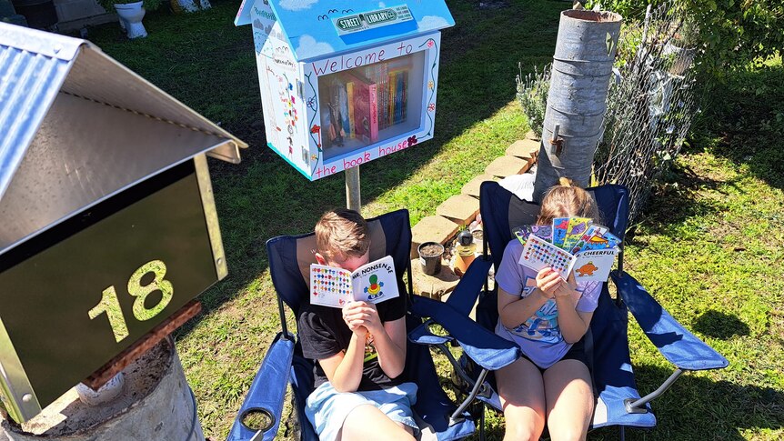 Two kids sitting in camp chairs hold books up to their faces 