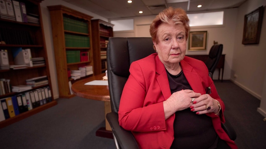 A woman with orange and white hair sits in an office hair in a room with bookcases