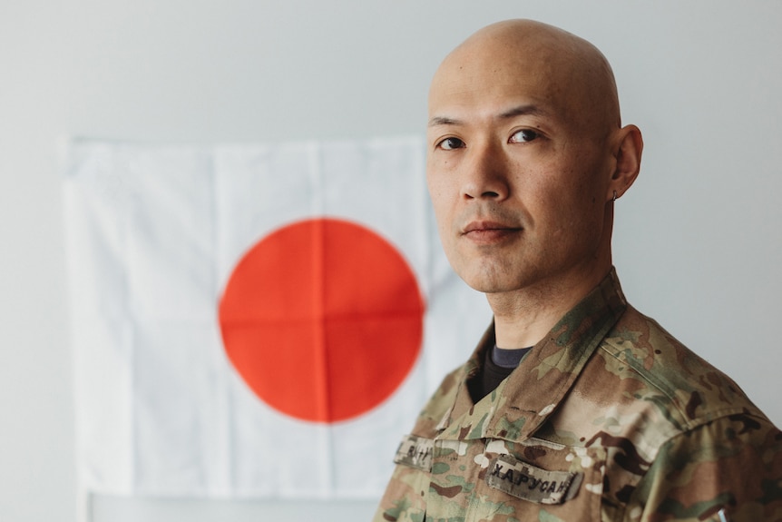 A bald man wearing cargo print military uniform stands in front of a Japanese flag