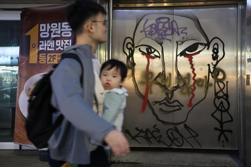 A man with a baby in a sling on his chest walk past line drawing art on a building window