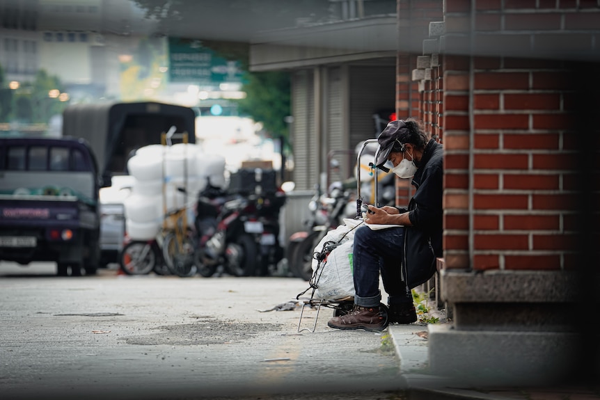 A man wearing a mask and cap sits next to garbage bags on the street.