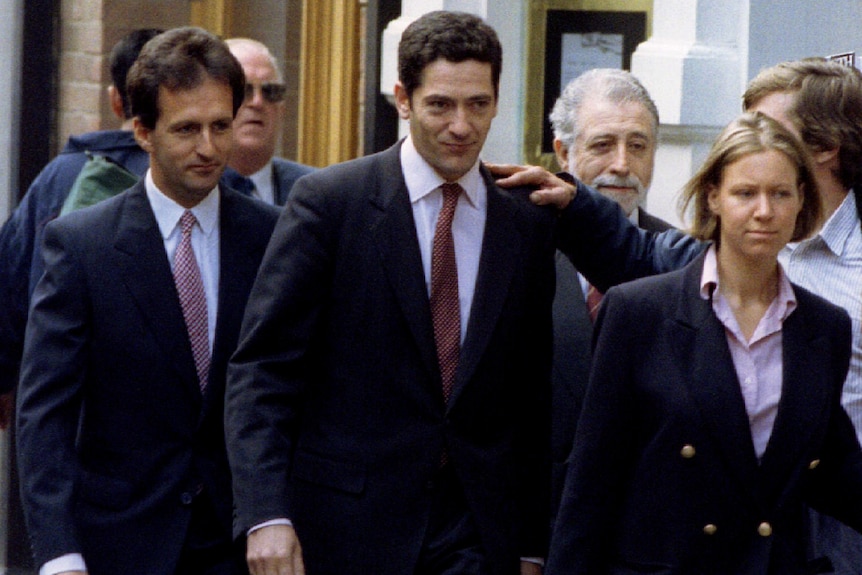 A man in a dark suit with a red tie accompanied by others walks in front of a court.