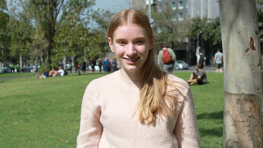 A teenage girl with long blonde hair, wearing a pale pink jumper, stands in a sunny park.