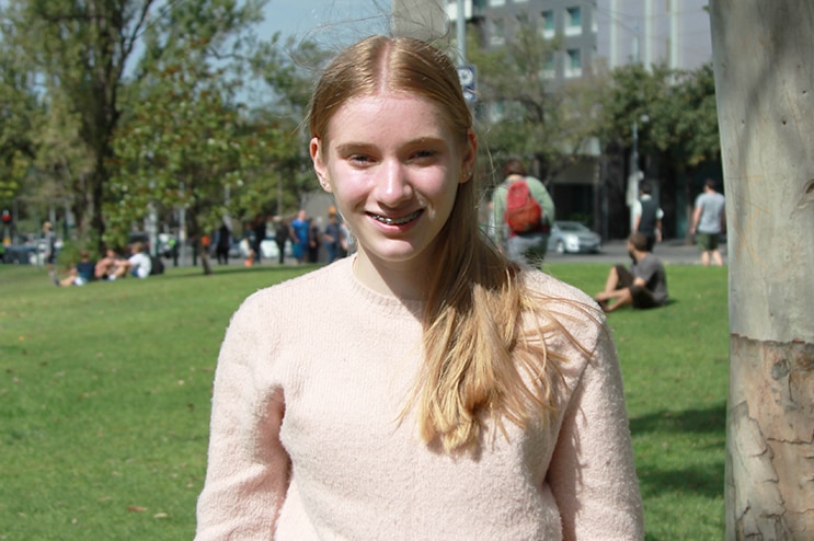 A teenage girl with long blonde hair, wearing a pale pink jumper, stands in a sunny park.