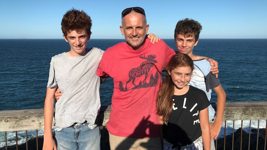 A father in a bright red shirt stands beside his three kids, with the ocean in the background. They're all smiling.