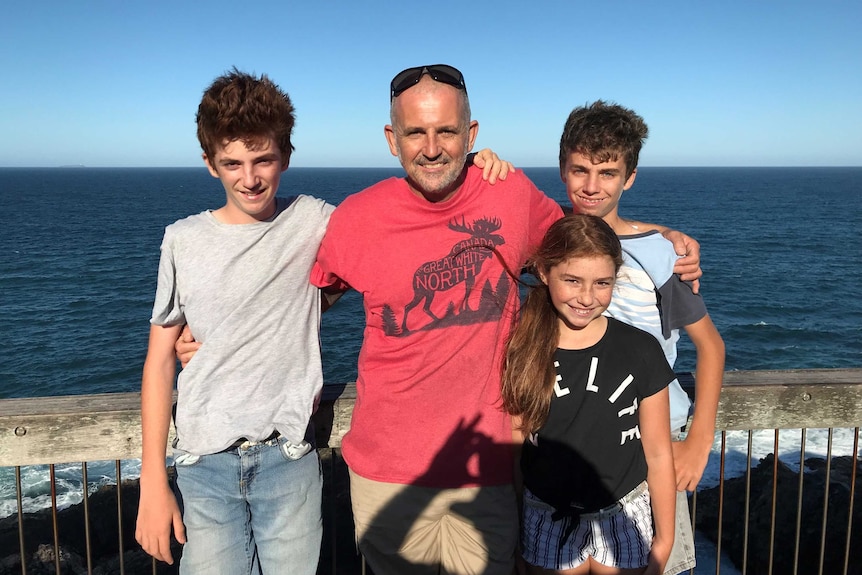 A father in a bright red shirt stands beside his three kids, with the ocean in the background. They're all smiling.