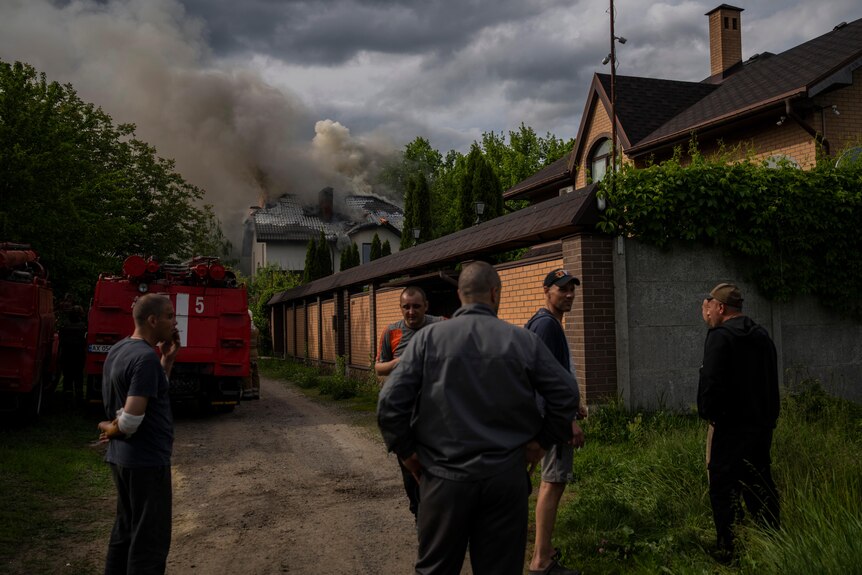 A group of men gather near a burning house in a residential area