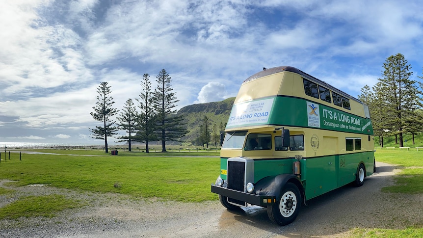 A double-decker bus parked by an ocean foreshore with mountains and pine trees in the background.