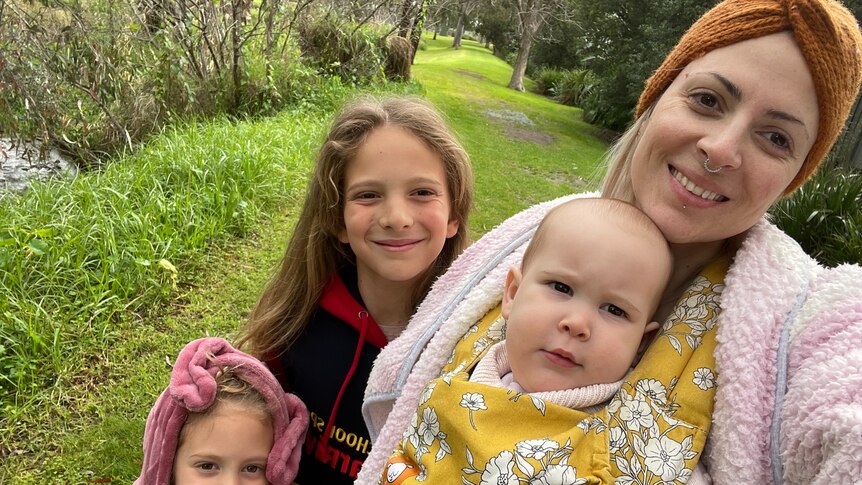 Mum in dressing gown smiles for a selfie with three young girls in a garden.